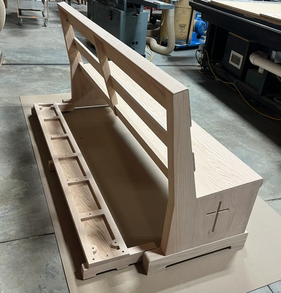 A fully assembled but unfinished custom wooden pew sitting on a workshop floor, showcasing its simple design and clean joinery.