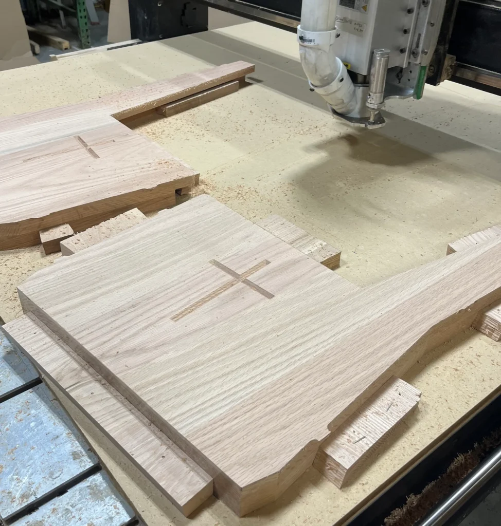 Detailed cross engravings on wooden pew ends resting on a CNC router bed in a woodshop.