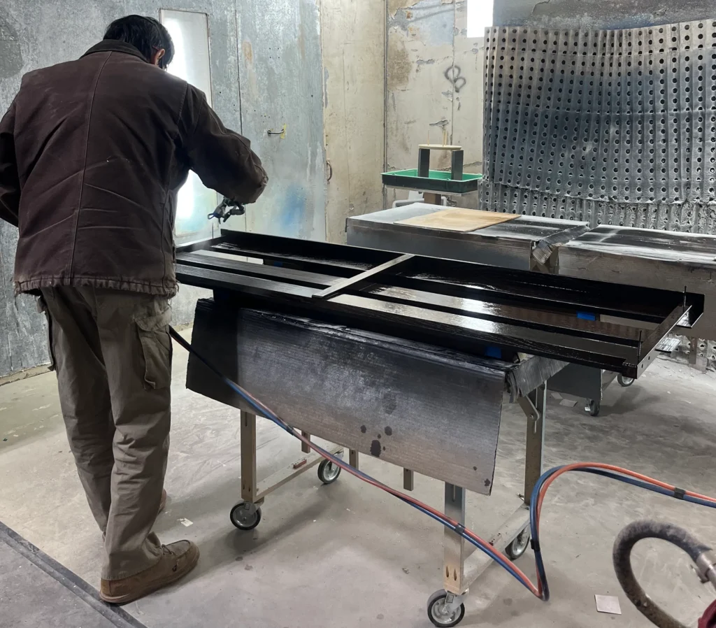 A professional woodworker wearing protective gear spraying a clear lacquer finish on wooden pew backs inside a spray booth.