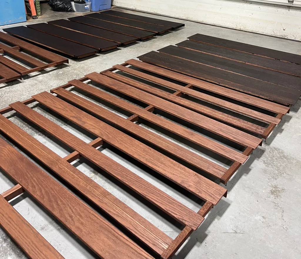 Rows of wooden pew backs and seats in a workshop during a multi-step staining process, showing the difference between the initial dye base and the final pigmented stain.